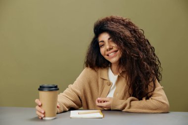 Sevimli, tatlı, bronzlaşmış kıvırcık, Latin bir kadın elinde kahve fincanı ile oturmuş zeytin yeşili arka planda not defteriyle kameraya bakıyor. Uzayı kopyala Mockup Banner. Serbest ofis.