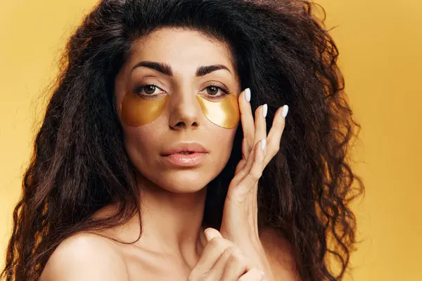 Curly pretty Latin woman in putting gold hydrogel mask on face for under-eye skin repair posing isolated over yellow background. Cosmetic product ad Natural beauty concept Studio closeup portrait