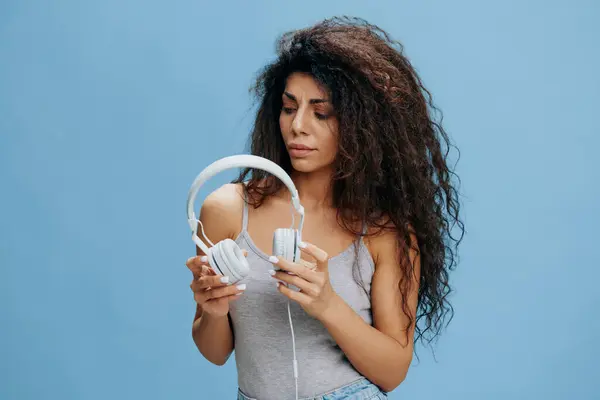 MUSIC STREAMING PLATFORM AD CONCEPT. Concentrated beautiful curly Latin female hold headphones and look on it, prepare to listen cool playlist. Cool offer for Phone pc ad karaoke. Over blue background