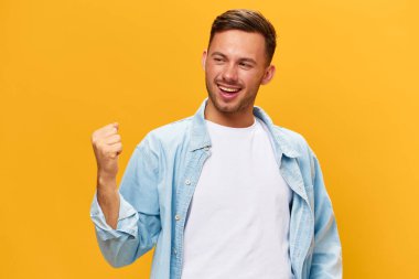 Şanslı gülümseyen, bronzlaşmış, mavi tişörtlü yakışıklı adam yumruğunu kaldır ve turuncu sarı stüdyo arka planında yalnız poz verdiğini söyle. Uzayın afiş modelini kopyala. İnsanların duyguları Yaşam tarzı konsepti