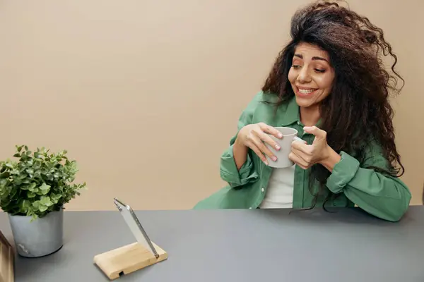 Enjoyed laughing happy tanned curly Latin lady hold cup of tea coffee sit at the table isolated over pastel beige background look at phone. Copy space Mockup Banner. Concept relax time