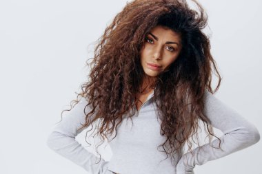 Afro kıvırcık saçlı, beyaz arka plan stüdyosunda yalnız poz veren, el ele kameraya bakan, kendine güvenen Latin bir kadının portresi. Moda ve kozmetik markaları için iyi bir teklif.