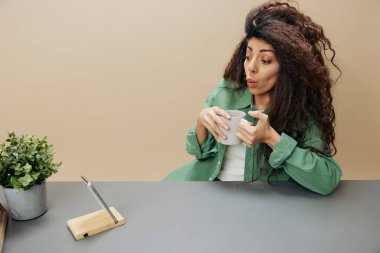 Heyecanlı, mutlu, bronzlaşmış, kıvırcık, Latin bir bayanın elinde çay fincanı tutarken pastel bej arka planda izole bir şekilde masaya oturup telefona bakması. Uzayı kopyala Mockup Banner. Rahatlama süresi kavramı