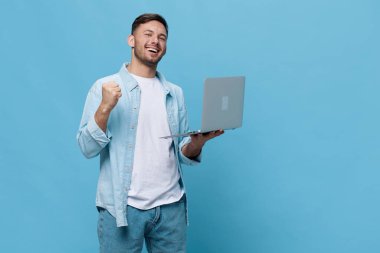 Keyifli, bronzlaşmış, sıradan tişörtlü yakışıklı adam mavi stüdyo arka planında dizüstü bilgisayarla başarılı bir galibiyete seviniyor. Uzayın afiş modelini kopyala. Elektronik tamir bilişim konsepti