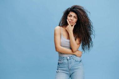 Elleri yanağında afro saçlı düşünceli, güzel Latin kadın, kameraya bak. Moda. Stüdyo çekimi mavi gökyüzü arka planında. Boşluğu kopyala, tasarım reklamı için boş yer