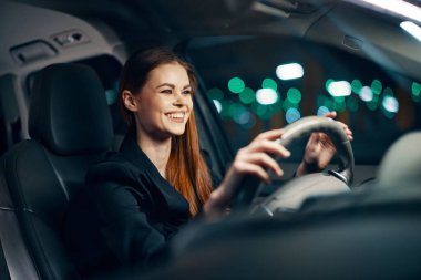 Siyah tişörtlü hoş bir kadının yatay fotoğrafı. Gece direksiyonda otururken araba sürüyor. Yüzünde hoş bir gülümsemeyle. Yüksek kalite fotoğraf