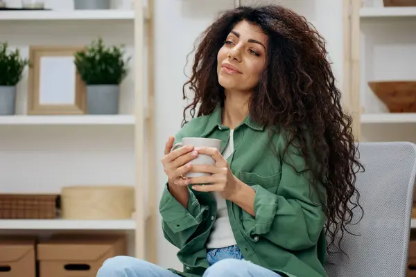 Cute smiling enjoyed pretty tanned curly Latin lady look with love aside drink tea coffee in casual shirt sit on chair in home modern interior background. Copy space Banner. Nostalgia concept