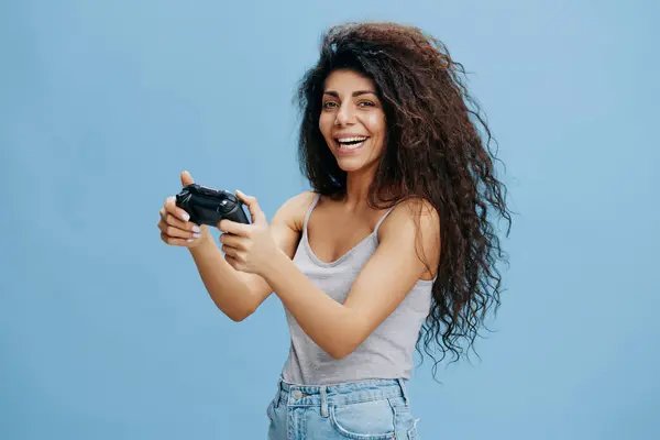 ONLINE GAME AD CONCEPT. Overjoyed beautiful curly Latin lady hold game pad in hands, ready to play cool RPG, smile, look at camera. Gaming accessories brands ad. Studio shoot isolated blue background