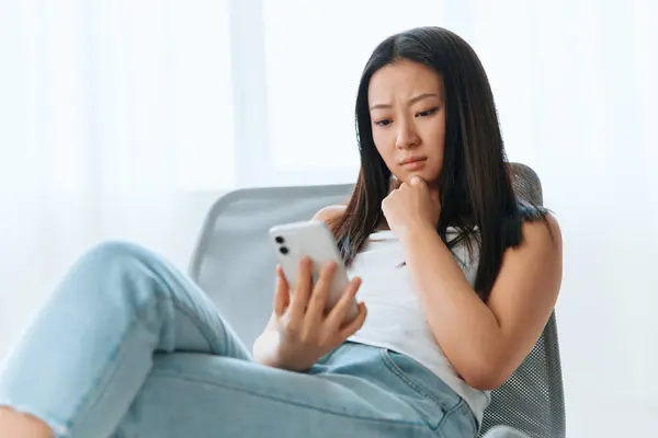 Upset confused tanned lovely young Asian woman read shocked message using phone at home interior living room. Distance communication, Remote work, Social media concept. Cool offer Banner Wide angle
