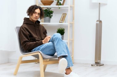 Kıvırcık saçlı bir adam, yaşam tarzında bir sandalyede oturan bir not defterine yazıyor. Yüksek kalite fotoğraf