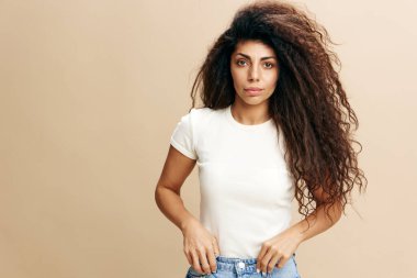 FASHION Önerisi. Seksi kıvırcık Arap kadın kameraya bakıyor, kot pantolonla el ele tutuşuyor. Moda markaları için fotokopi alanı, reklam için bedava yer. Bej arkaplanlı stüdyo çekimi