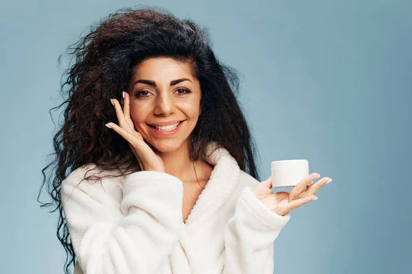 Smiling happy laughing curly Latin lady in bathrobe looking at camera hold jar of cream, touching cheek, posing isolated on pastel blue background. Organic cosmetic product ad concept. Natural beauty