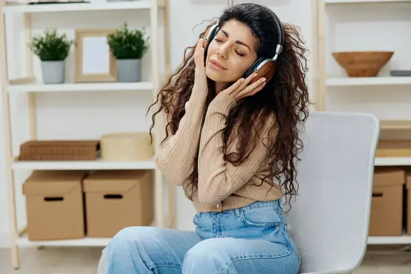 Happy relaxed pleasant tanned curly Latin lady with headphones listen cool playlist sit on chair in home modern interior close eyes. Copy space Mockup Banner. Concept sound stream. Relax in home
