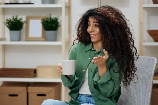 Happy laughing enjoyed pretty tanned curly Latin lady talking with friend hold cup of tea coffee in casual shirt sit on chair in home modern interior background look aside. Copy space Banner