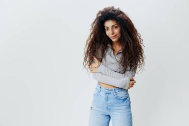 Modern tarz. Moda Teklifi. Kendine güveni tam kıvırcık kıvırcık, rahat giyinen, gülümseyen, sola bakan, katlanmış koluyla poz veren, izole beyaz arka planlı Latin bir kadın. Boş yer reklamını kopyala. 