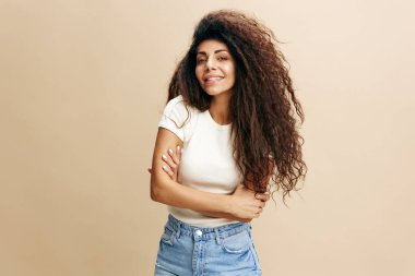 Kadın portresi. Happy, kolları katlanmış, kameraya gülümseyen beyaz tişörtlü, bej renkli stüdyo arka planında yalnız poz veren Latin kadınlardan hoşlanırdı. Olumlu kadın modeli gülüyor. 