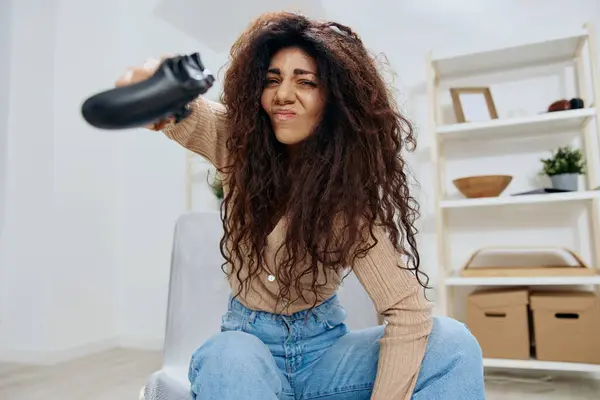 Irritated angry pretty tanned curly Latin lady lose game throw joystick gamepad at camera sit on chair in home modern interior. Copy space Mockup Banner. Concept gamer. People emotions concept