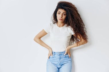 Beyaz tişörtlü zayıf, kendine güvenen, kıvırcık saçlı, beyaz arka plan stüdyosunda yalnız poz veren belinde el ele kameraya bakan Latin bir kadın. Moda ve kozmetik markaları için iyi bir teklif.