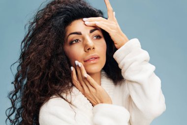 Bornozlu sevimli kıvırcık Latin bayan kusursuz tenine dokunuyor, besleyici bir maske takıyor, pastel mavi arka planda yalnız poz veriyor. Yüz kremi reklamı konsepti. Boşluğu kopyala