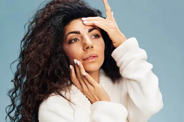 Bornozlu sevimli kıvırcık Latin bayan kusursuz tenine dokunuyor, besleyici bir maske takıyor, pastel mavi arka planda yalnız poz veriyor. Yüz kremi reklamı konsepti. Boşluğu kopyala