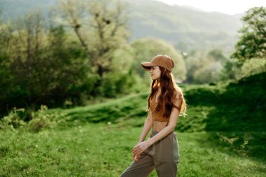 Spor giyim ve şapkalı güzel bir kadın yeşil bir doğal manzara üzerinde yürüyor ve güneş ışığında gülümsüyor. Yüksek kalite fotoğraf