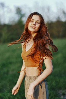 Doğada uzun kıvırcık saçlı, gün batımının tadını çıkaran ve kameraya bakarken gülümseyen bir kadın. Yüksek kalite fotoğraf