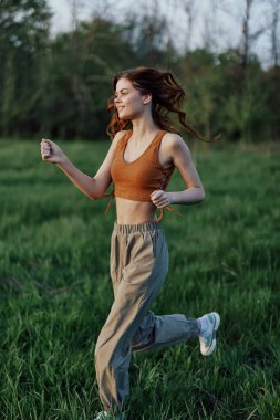 Atletik bir kadın yeşil pantolon ve spor ayakkabılarla çimenlerde koşar ve yaz mevsimi için vücudunu gün batımında parkta eğitir. Yüksek kalite fotoğraf
