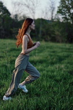 Uzun kızıl saçlı bir kadın spor yapıyor ve parktaki yeşil çimlerde eşofman ve spor ayakkabılarıyla koşuyor. Yüksek kalite fotoğraf