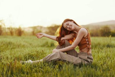 Bir kadın açık havada yeşil taze çimenlerin üzerinde oturur ve gün batımında mutlu bir şekilde güler. Uçan kızıl saçları rüzgarda dalgalanır. Yüksek kalite fotoğraf