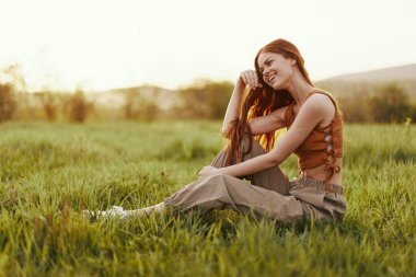 Bir kadın açık havada yeşil taze çimenlerin üzerinde oturur ve gün batımında mutlu bir şekilde güler. Uçan kızıl saçları rüzgarda dalgalanır. Yüksek kalite fotoğraf
