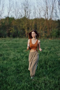 Atletik yapılı bir kadın yeşil çimlerin üzerinde koşuyor, gevşek, uçan saçlarıyla ve gün batımına doğru egzersiz yapıyor. Yüksek kalite fotoğraf