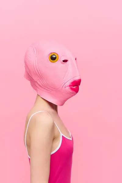A woman in a pink fish head mask stands in profile against a pink background and looks into the camera with one yellow eye, a crazy conceptual Halloween costume. High quality photo