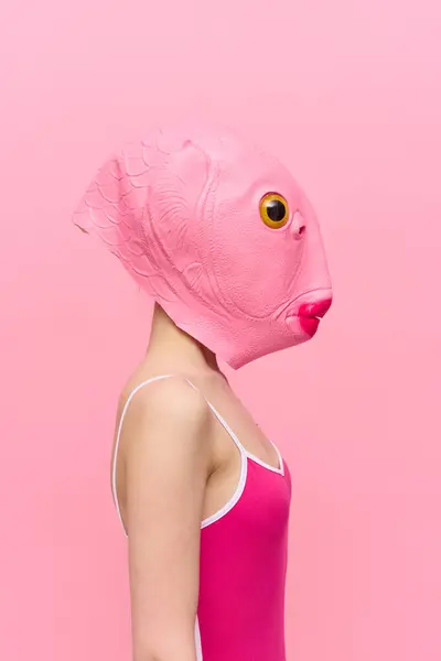 A woman in a pink fish head mask stands in profile against a pink background and looks into the camera with one yellow eye, a crazy conceptual Halloween costume. High quality photo