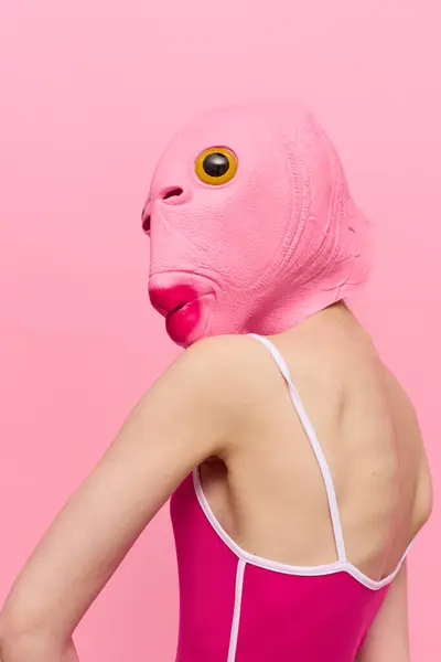 Skinny woman in a pink Halloween costume with a fish head on her face poses funny against a pink background and looks at the camera, art concept photo. High quality photo