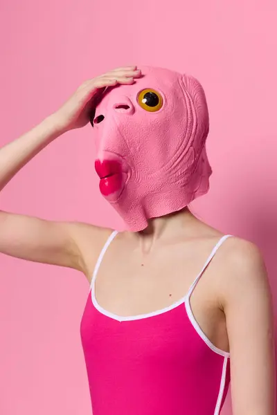 Woman with fish head mask on pink is holding her head in surprise, modern art photo on pink background. High quality photo
