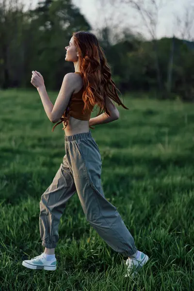 長い赤い髪の女性は スウェットパンツやスニーカーで公園の緑の草の上を走ります 高品質の写真 — ストック写真