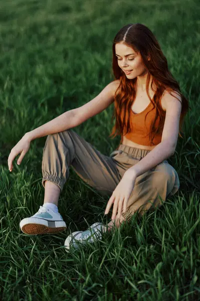 A beautiful skinny woman sits on the grass in casual clothes and relaxes from exercising in the sunset light. High quality photo