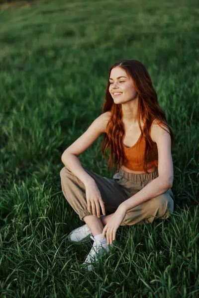 A beautiful skinny woman sits on the grass in casual clothes and relaxes from exercising in the sunset light. High quality photo