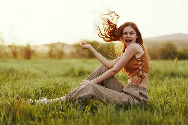 A woman sits on the green young fresh grass in the open air and laughs happily in the sunset light, her flying red hair fluttering in the wind. High quality photo