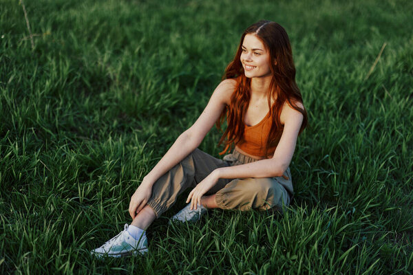 A beautiful skinny woman sits on the grass in casual clothes and relaxes from exercising in the sunset light. High quality photo