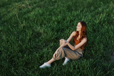 Uzun, kıvırcık kızıl saçlı güzel bir kadın yazın gün batımında yeşil çimlerin üzerinde oturur. Yüksek kalite fotoğraf