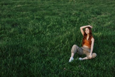 Rahat giysiler içindeki bir kız parkta yürüdükten sonra çimlere oturdu meditasyon yapmak ve gün ışığında dinlenmek için. Yüksek kalite fotoğraf