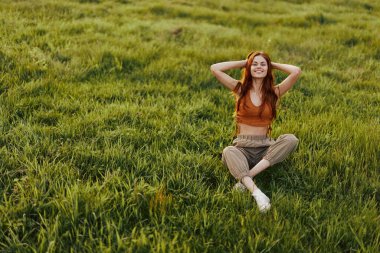 Kızıl saçlı bir kadın ellerini başının arkasına koyup çimlerin üzerinde oturur ve gün batımında gülümser. Sağlıklı dinlenme ve sağlık için meditasyon kavramı. Yüksek kalite fotoğraf