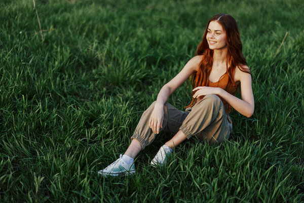 A beautiful skinny woman sits on the grass in casual clothes and relaxes from exercising in the sunset light. High quality photo