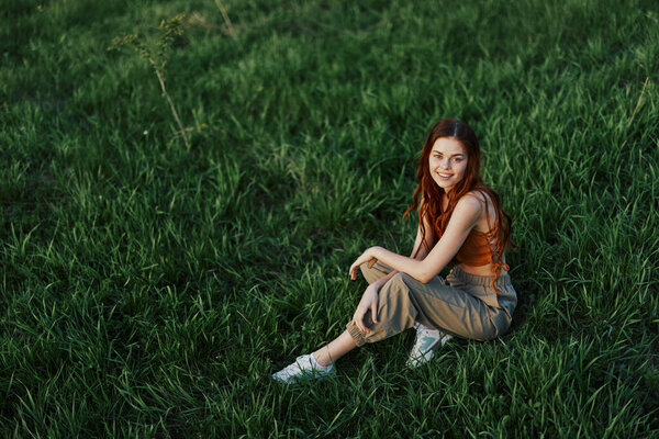 A beautiful woman with long, curly red hair sits in the park in the summer on the green grass in the sunset light. High quality photo