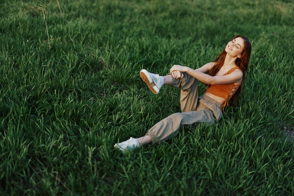 The redheaded woman sits in the park on the green grass wearing an orange top, green pants, and sneakers and looks out at the setting summer sun. High quality photo