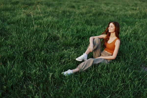 The redheaded woman sits in the park on the green grass wearing an orange top, green pants, and sneakers and looks out at the setting summer sun. High quality photo