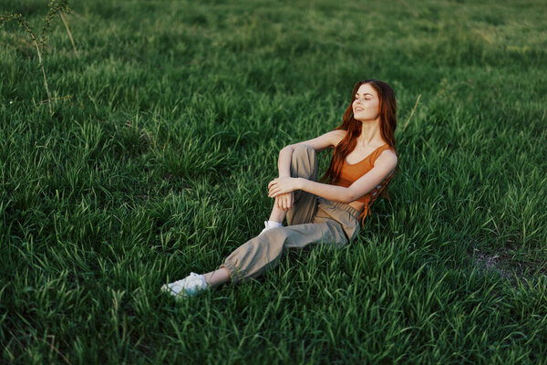 The redheaded woman sits in the park on the green grass wearing an orange top, green pants, and sneakers and looks out at the setting summer sun. High quality photo