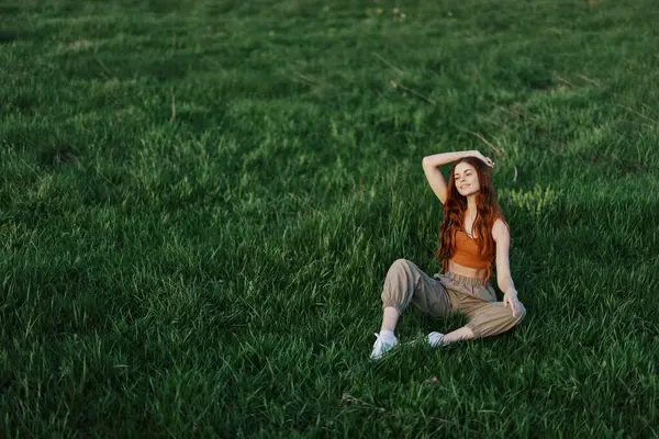 A girl in comfortable clothes sat on the grass after a walk in the park to meditate and rest in the sunlight. High quality photo