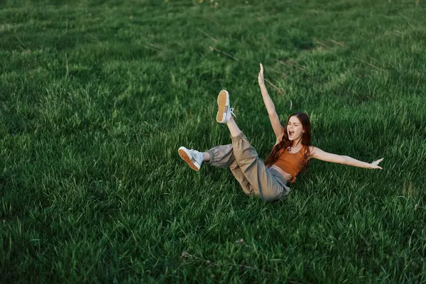 A young woman playing games in the park on the green grass spreading her arms and legs in different directions falling and smiling in the summer sunlight. High quality photo